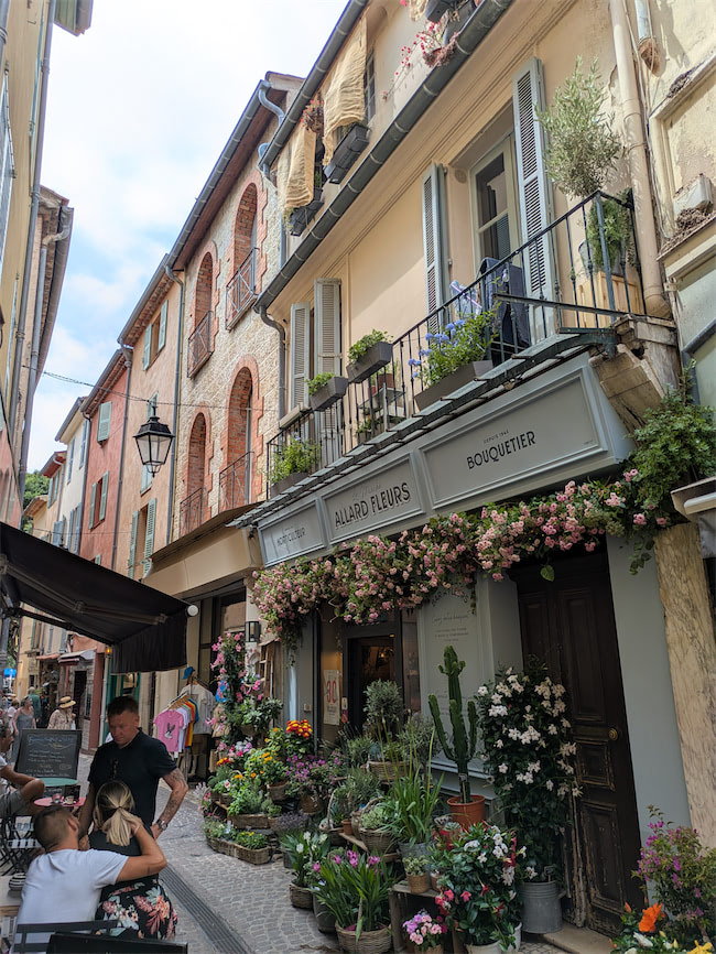 A narrow European street lined with colorful buildings and balconies, featuring a flower shop with abundant plants and blooms outside. People stroll and sit at a café, creating a lively, charming atmosphere.