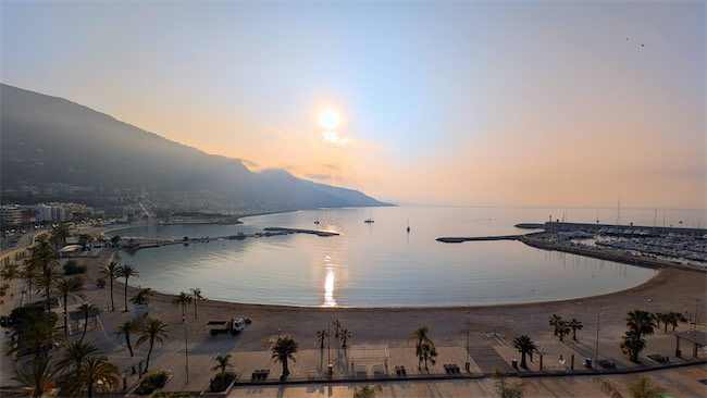 A calm bay with palm trees and sandy beach, bordered by a promenade. The sun is low over mountains, casting a golden light across the tranquil sea and marina, with boats visible in the water.