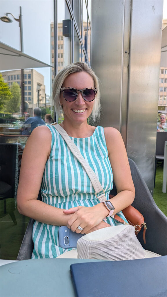 A woman with blonde hair, wearing sunglasses and a blue-and-white striped dress, sits and smiles at an outdoor cafe table. She has a white crossbody bag and a smartwatch. Buildings and greenery are visible in the background.