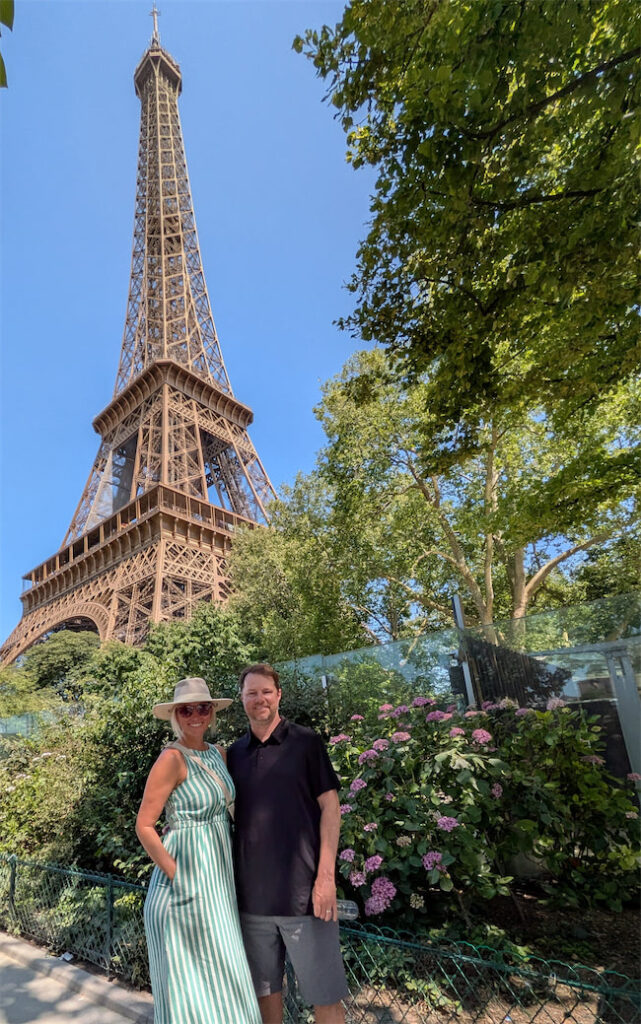 A smiling couple poses in front of the Eiffel Tower on a sunny day, surrounded by green trees and blooming flowers. The woman wears a striped dress and hat, while the man wears a black shirt and shorts.