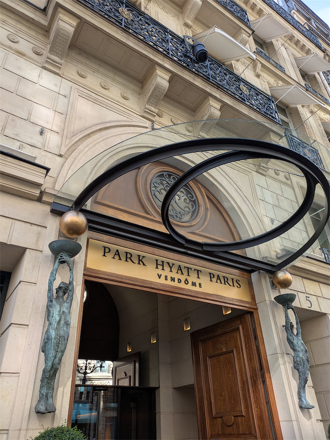 Entrance of Park Hyatt Paris Vendôme, featuring ornate stonework, two bronze sculptures holding globes, a wooden door, and a glass canopy with metal rings above the entrance.