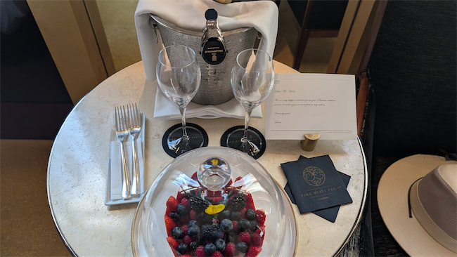 A round table with a plate of mixed berries under a glass dome, two empty wine glasses, a bottle of champagne in an ice bucket, folded napkins with cutlery, a card, and a hat placed nearby.