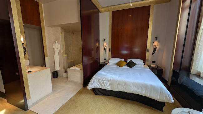 A modern hotel room with a large bed, white linens, dark wood accents, and gold trim. An adjacent open bathroom area has a robe hanging, a shower, and a vanity with a mirror. Natural light enters through a window.