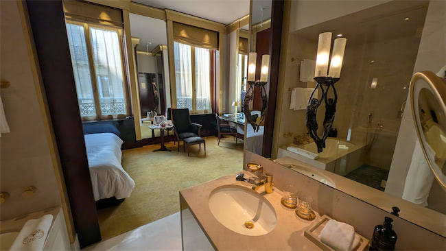 Elegant hotel room with a modern bathroom in the foreground, featuring a large mirror, sink, and toiletries. The bedroom area has a cozy bed, chairs, a desk, and tall windows letting in natural light.