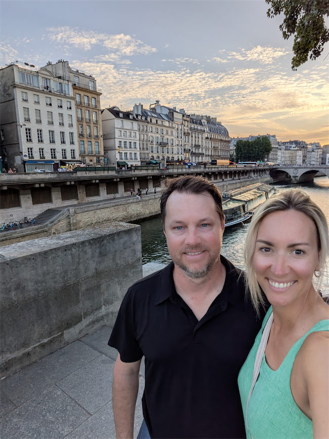 A smiling couple takes a selfie near a river with historic buildings and a bridge in the background under a sunset sky.