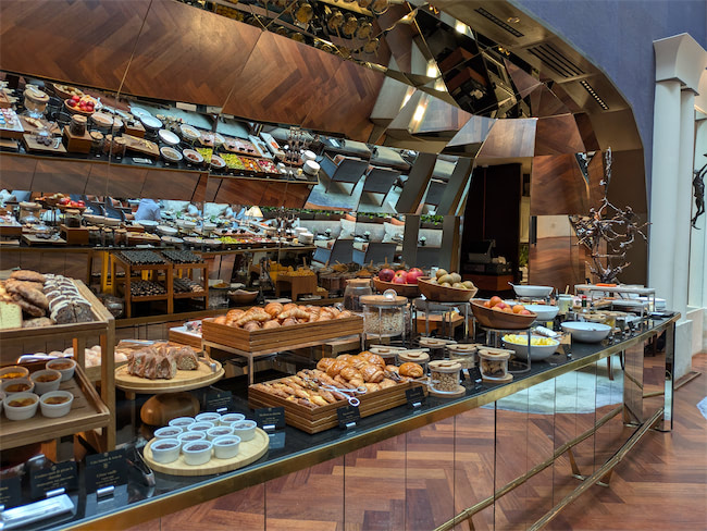 A luxurious breakfast buffet features pastries, bread, fruit, yogurt, and spreads neatly arranged on wooden trays and bowls, with a mirrored wall reflecting the elegant display.