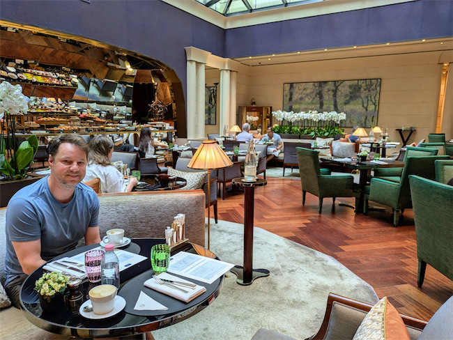 A man sits at a table with coffee and pastries in an elegant, well-lit café with plush seating, wooden floors, and large windows. Other patrons dine in the background.