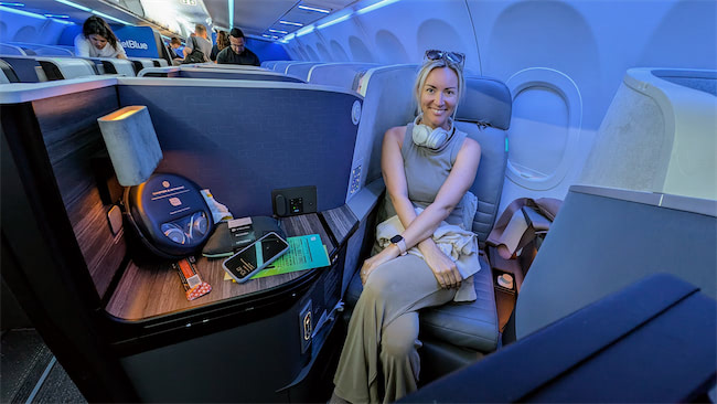 A woman sits comfortably in a spacious, modern airplane business class seat with a table holding travel items, under blue ambient lighting. Other passengers are seen in the background.