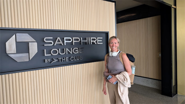 A woman with headphones around her neck and a backpack stands smiling in front of a sign that reads Sapphire Lounge by The Club inside an airport or lounge area.