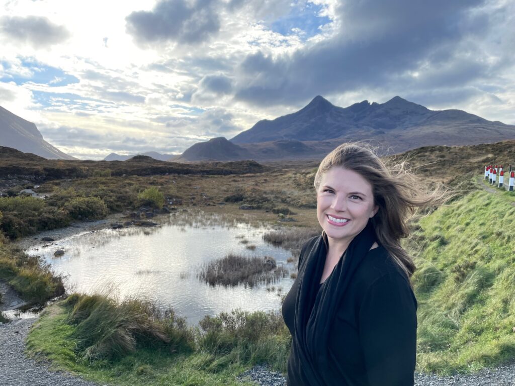 A woman with long brown hair in a black coat smiles at the camera, standing near a small pond with mountains and a cloudy sky in the background.