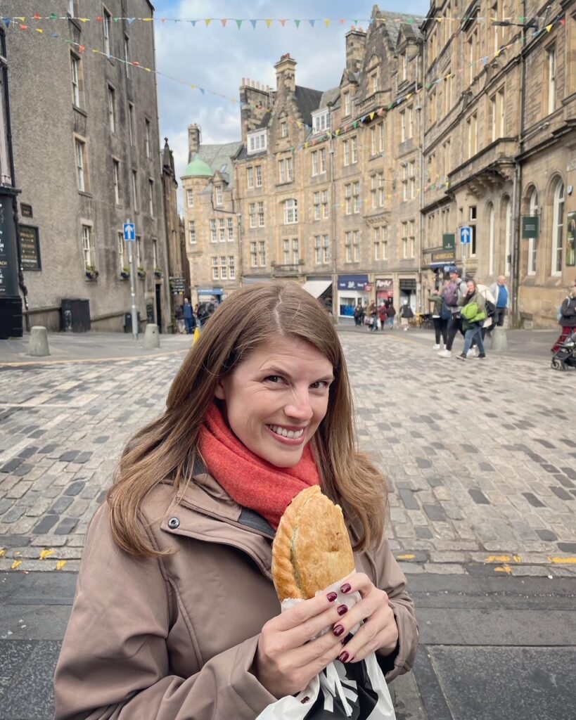 A woman in a brown coat and red scarf smiles while holding a pastry on a cobblestone street, with historic stone buildings and people walking in the background.