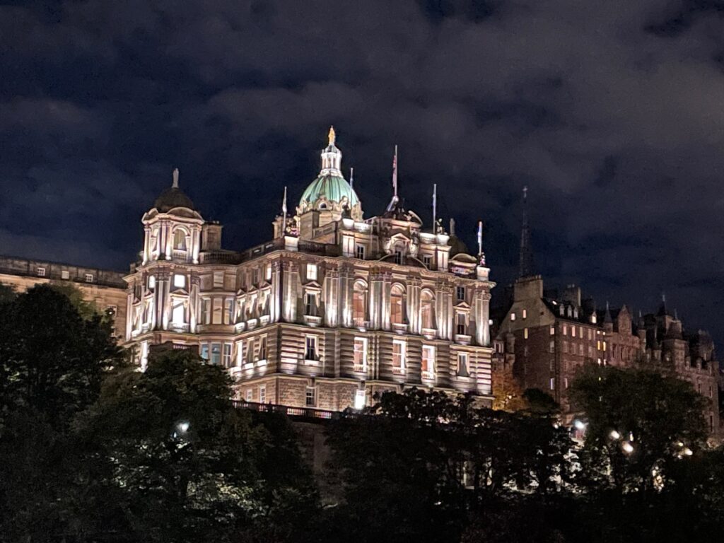 A large historic building with a green dome and ornate architecture is illuminated at night, with trees in the foreground and a dark, cloudy sky above.