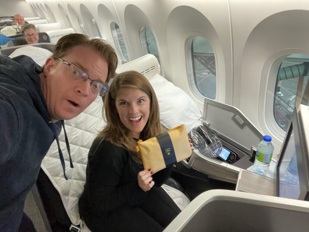 A man and woman smiling in a business class airplane cabin; the woman holds a yellow amenity kit, and the seat has a water bottle, headphones, and a large pillow. Plane windows and another passenger are visible in the background.