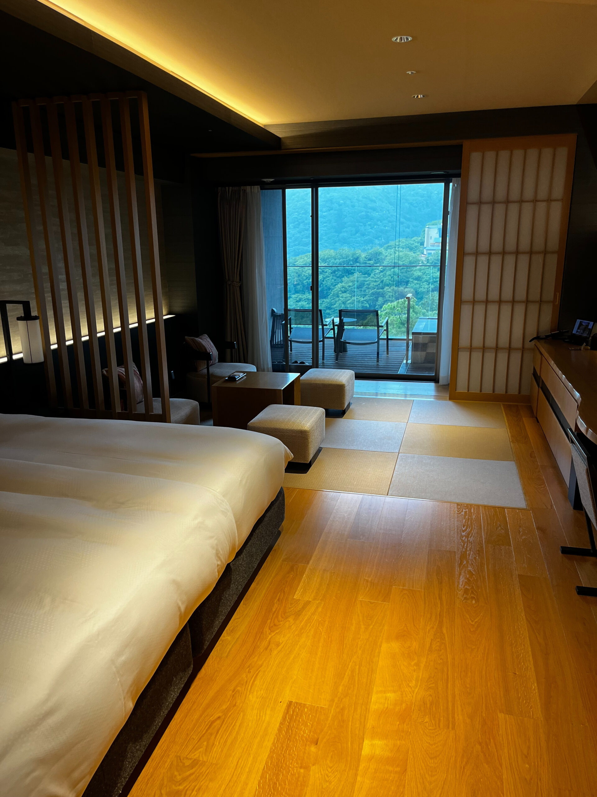 A modern, warmly lit hotel room with a large bed, seating area, and sliding glass doors opening to a balcony with mountain views. The room features wood and paper screen accents, blending Japanese and contemporary design.