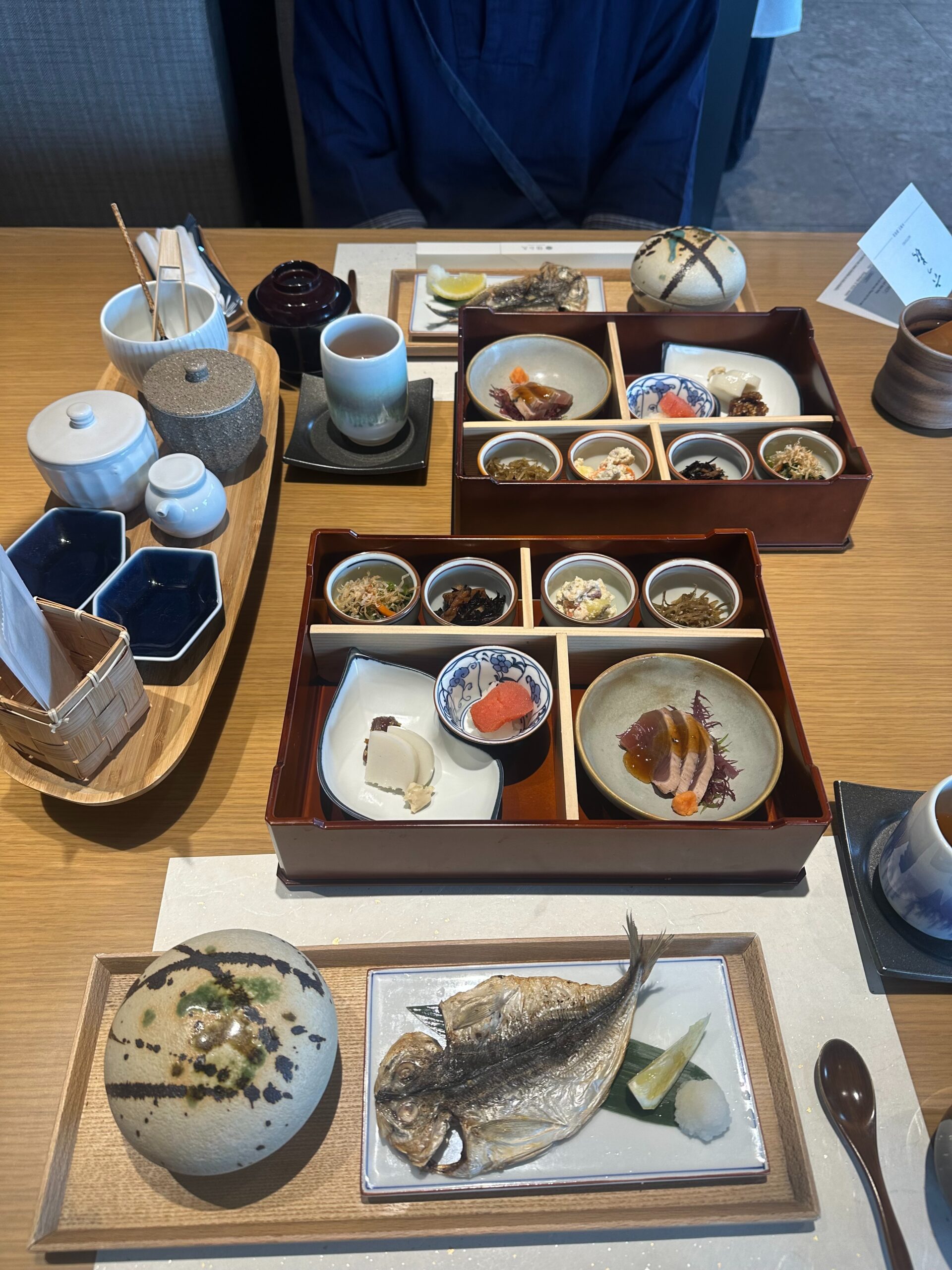 A traditional Japanese meal set on a wooden table, featuring grilled fish, assorted small dishes, rice in covered bowls, tea, and condiments, all neatly arranged in trays and plates.