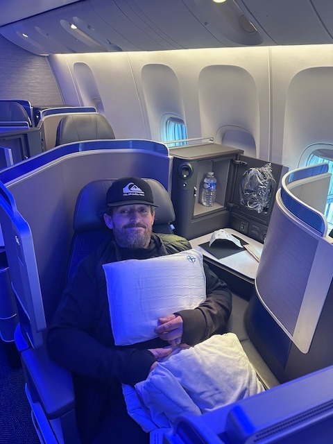 A man wearing a baseball cap sits comfortably in a spacious airplane business class seat, holding a pillow and blanket, with a water bottle and headphones nearby. Blue ambient lighting fills the cabin.