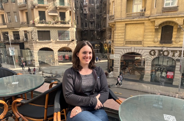 A woman with brown hair sits at an outdoor café table on a balcony, smiling, with a city street and yellow historic buildings visible in the background.