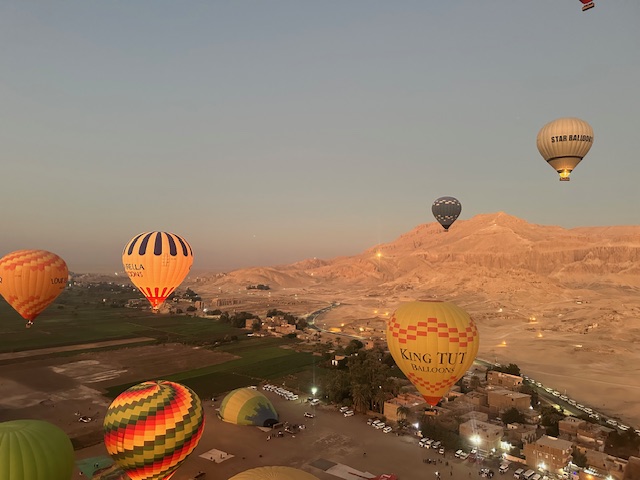 Hot air balloons of various colors and patterns float above fields and ancient ruins at sunrise, with a desert mountain landscape in the background. Some balloons have names like King Tut Balloon visible.