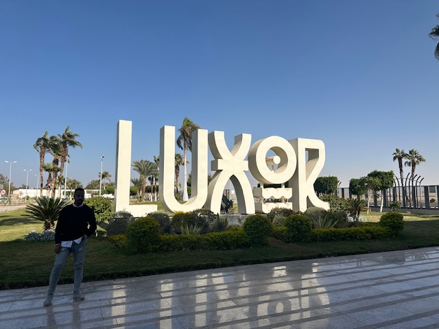 A man stands in front of a large white LUXOR sign surrounded by greenery and palm trees, with a clear blue sky in the background.