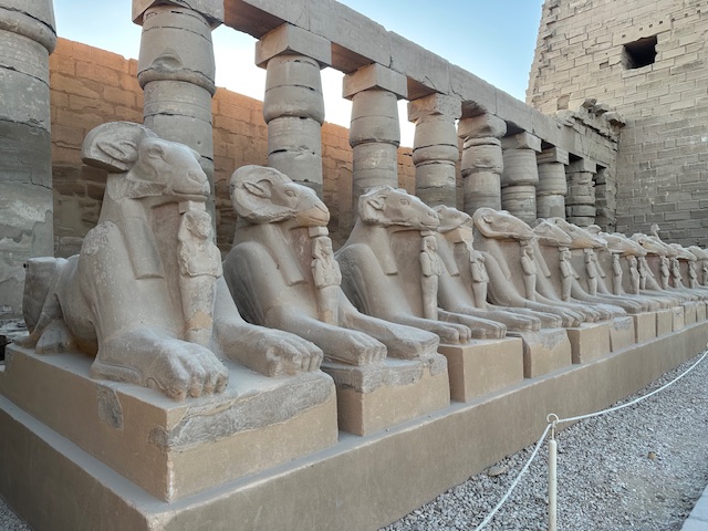 A row of ancient Egyptian ram-headed sphinx statues lines a stone pathway in front of tall, weathered columns at an archaeological site.