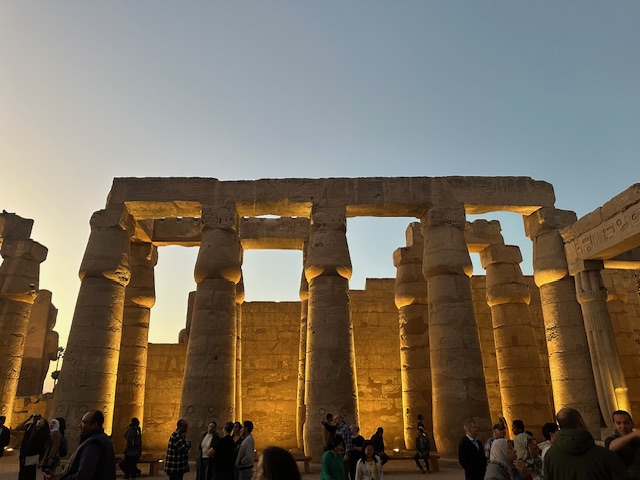 Ancient stone columns of an Egyptian temple are illuminated at sunset, with a clear sky above. Visitors walk and stand around the site, casting shadows on the ground.