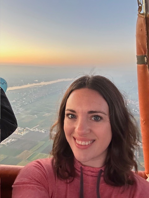 A woman with brown hair smiles for a selfie inside a hot air balloon, with a scenic view of fields, a river, and a colorful sky at sunrise or sunset in the background.