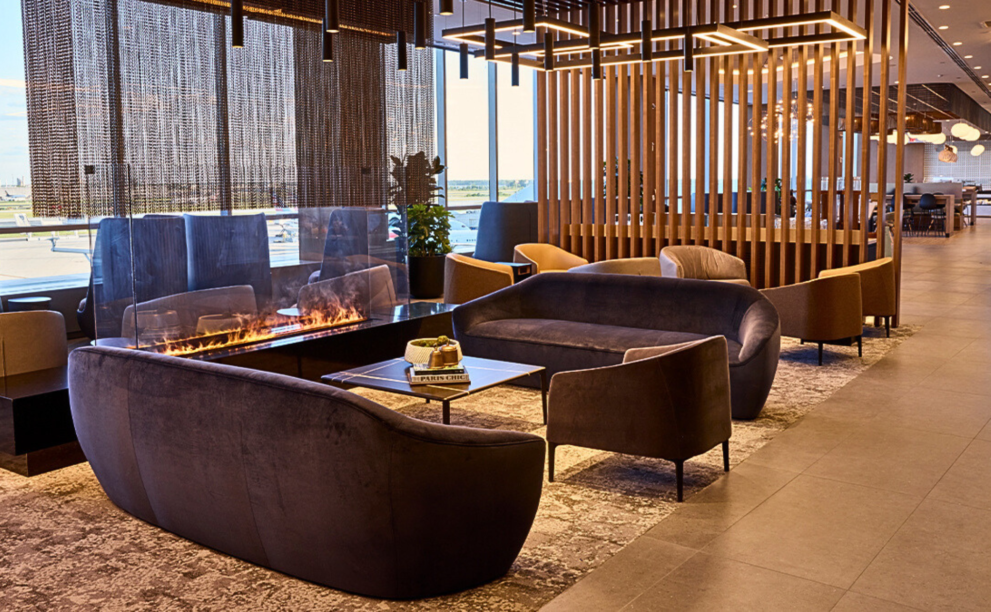A modern airport lounge with velvet sofas and armchairs around a rectangular glass fireplace, floor-to-ceiling windows, wooden slat dividers, and a view of the runway outside.