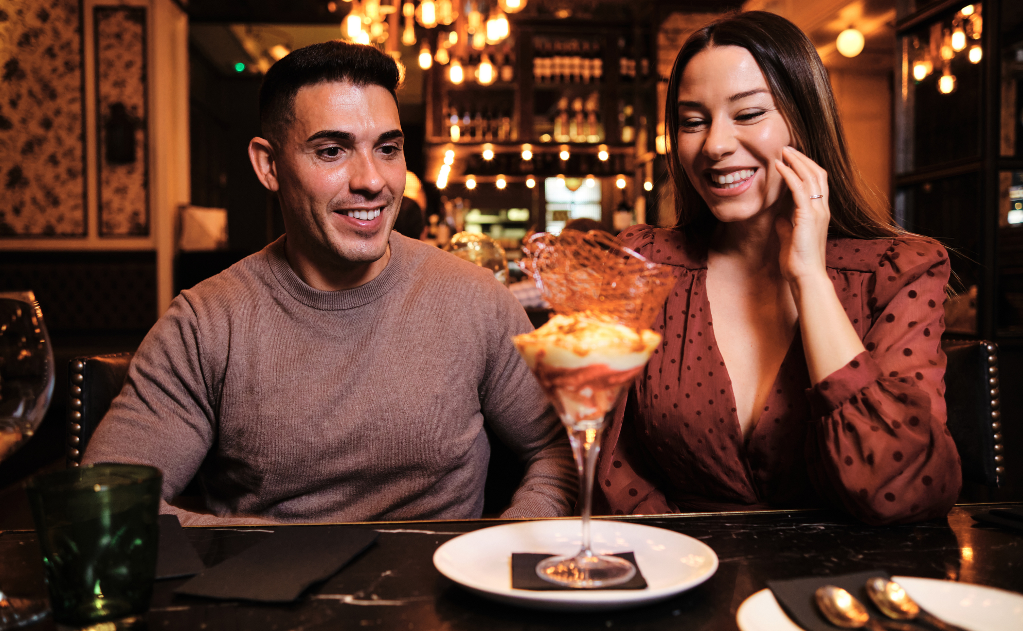 A smiling man and woman sit together at a restaurant table, looking excitedly at a fancy dessert in a glass. Warm hanging lights and elegant decor create a cozy, romantic atmosphere.