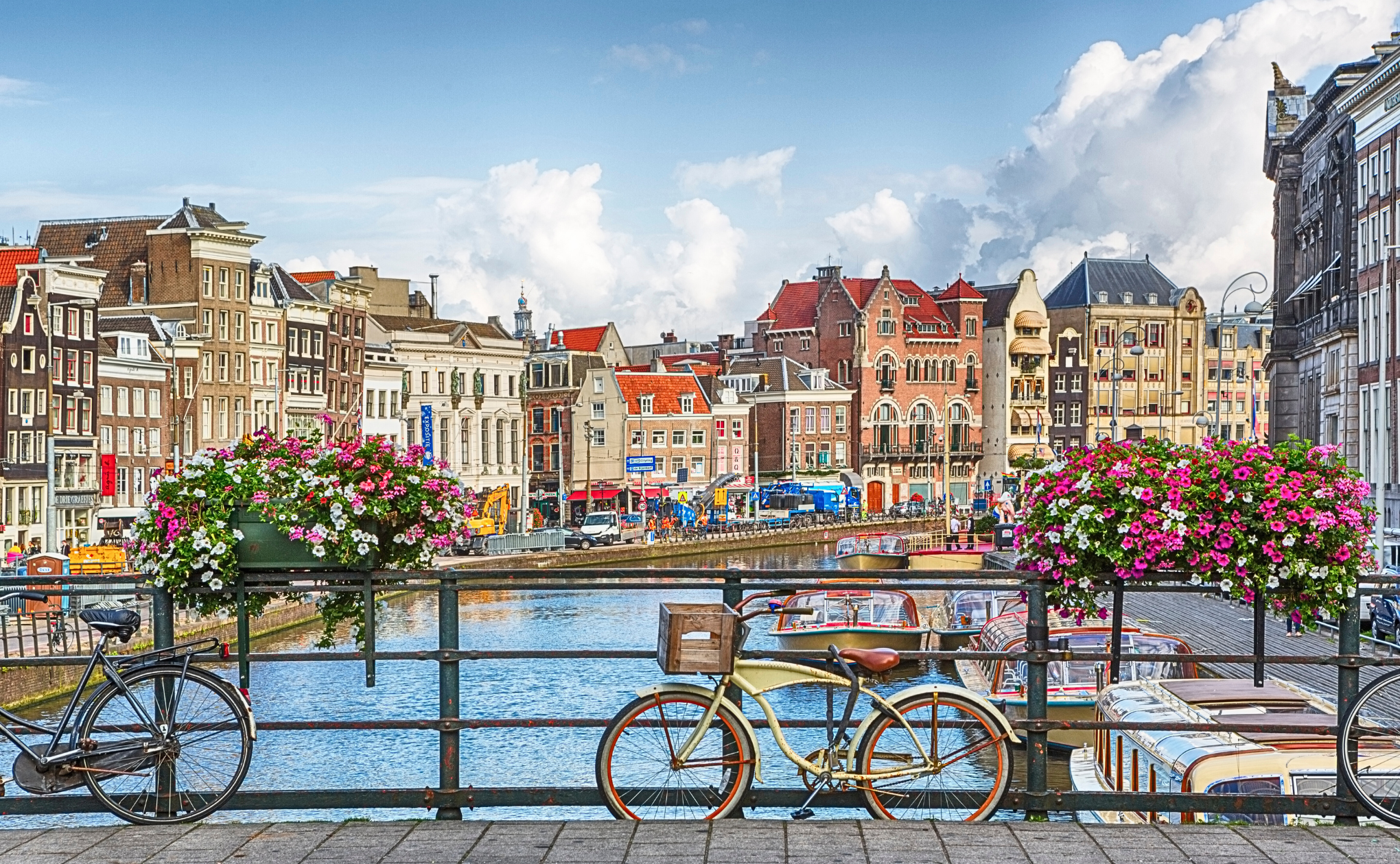 Bicycles parked on a canal bridge in Amsterdam, with colorful flowers on the railings, historic buildings lining the water, and boats docked along the canal under a partly cloudy sky.