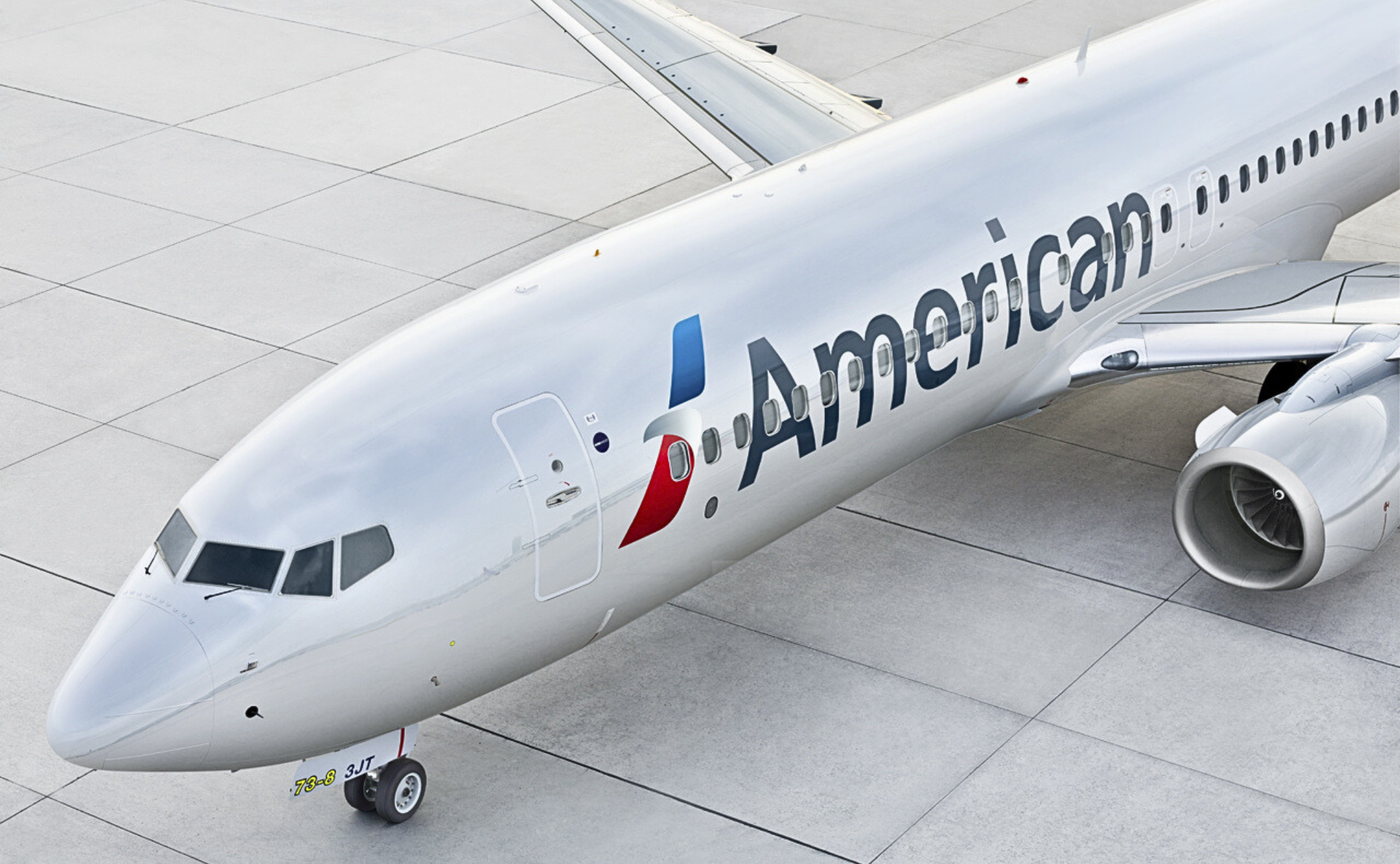 An American Airlines jet is parked on a concrete airport surface, viewed from above and slightly to the side, showing the front half of the aircraft with the company logo and part of the right engine visible.