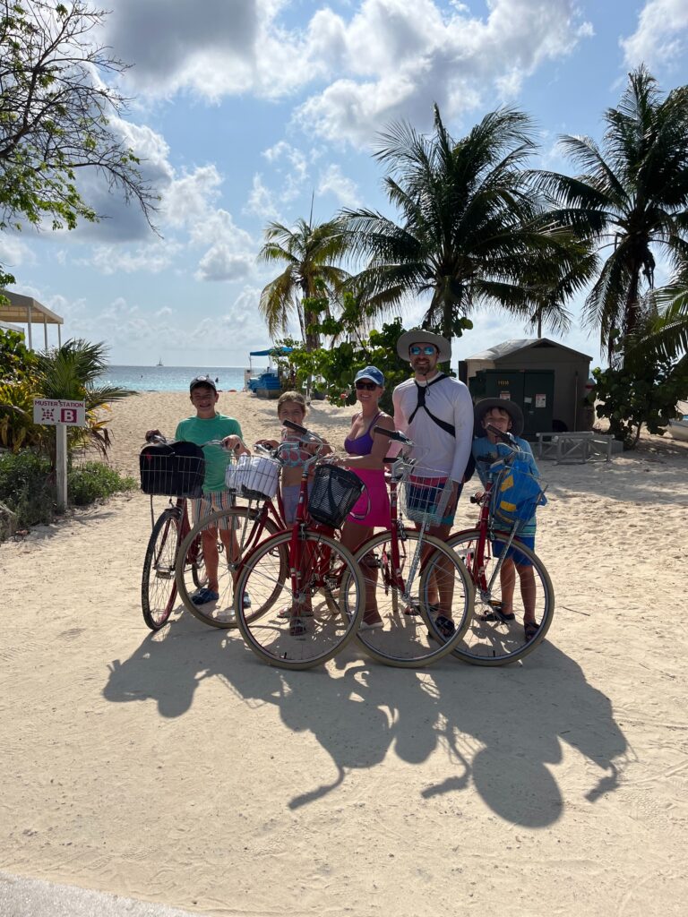 A family of five stands with their bicycles on a sandy path near the beach, surrounded by palm trees. The ocean and a blue sky with scattered clouds are in the background.