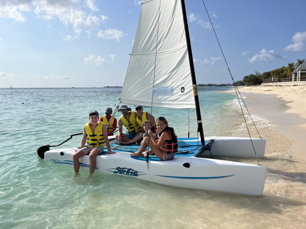 A group of six people wearing life jackets sit on a white Hobie catamaran at the edge of a sandy beach. The boat is in shallow, clear water under a sunny sky with light clouds.