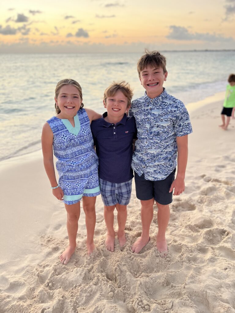 Three smiling children stand barefoot on a sandy beach at sunset, with the ocean and a partly cloudy sky in the background. Two boys and a girl, all wearing blue, pose together near the water’s edge.