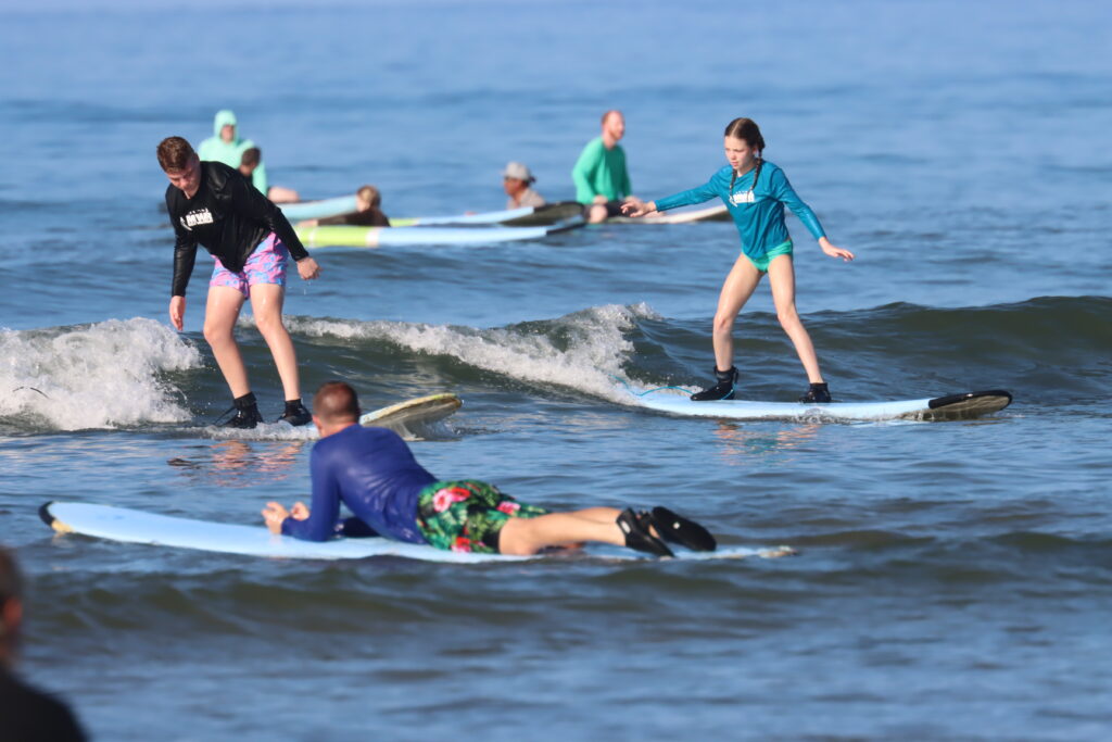 Several people are surfing small waves in the ocean; two girls are standing on surfboards while a man in a blue shirt and floral shorts lies on his board in the foreground. Other surfers are in the background.