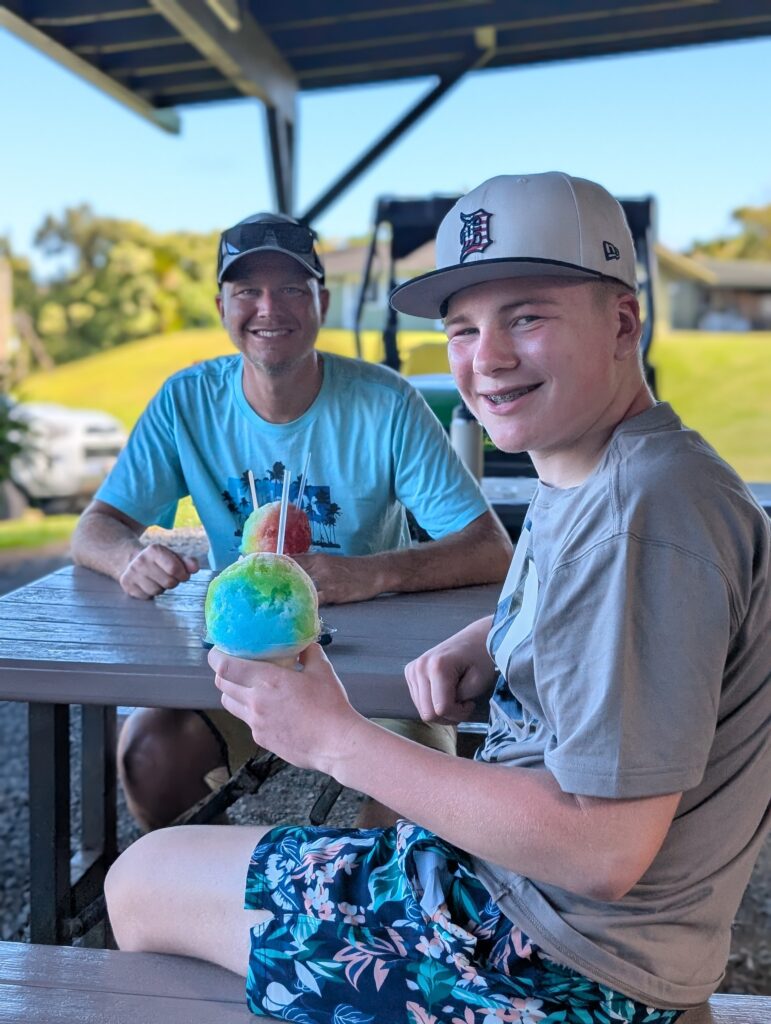 Two smiling people sit at a picnic table outdoors. The person in front holds a colorful shaved ice treat. Both wear casual clothes and baseball caps. The background shows green grass, trees, and a bright sunny day.