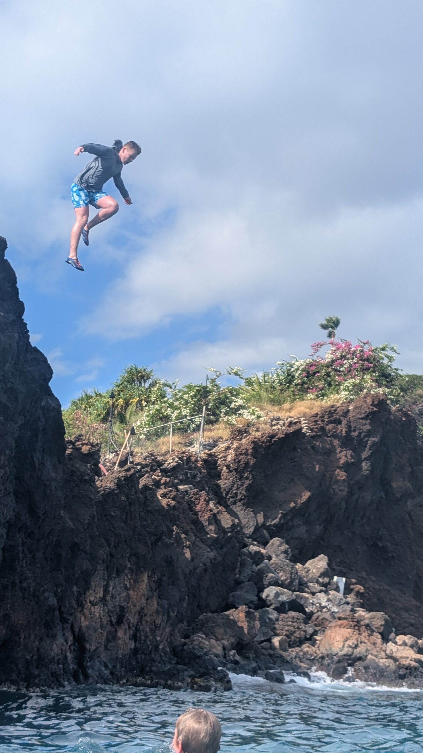 A person in a dark shirt and blue shorts is captured mid-air while jumping off a rocky cliff into the water below, with greenery and a partly cloudy sky in the background.