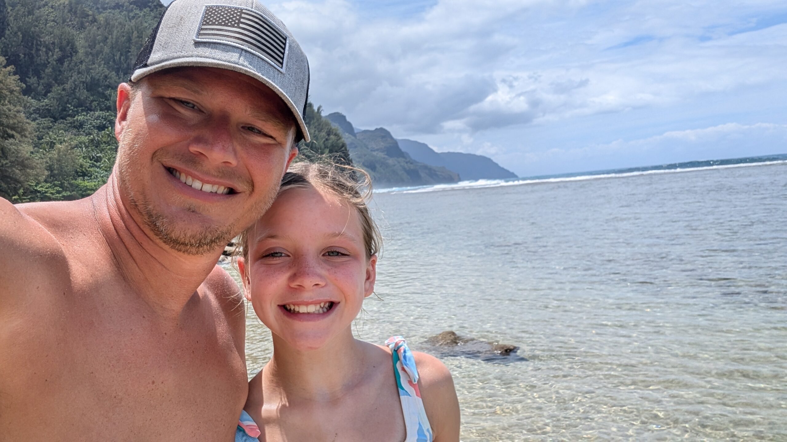 A man and a young girl smiling for a selfie on a rocky beach with clear water, lush green mountains, and a partly cloudy sky in the background.