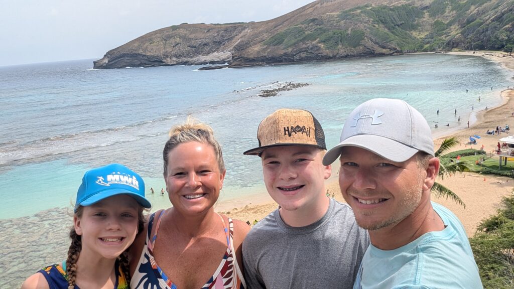 A smiling family of four poses for a selfie on a beach with clear blue water, rocky shoreline, and green hills in the background on a sunny day.