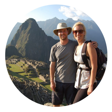 A man and woman smiling and posing together at Machu Picchu, with green mountains and ancient stone ruins in the background, under a clear sky.