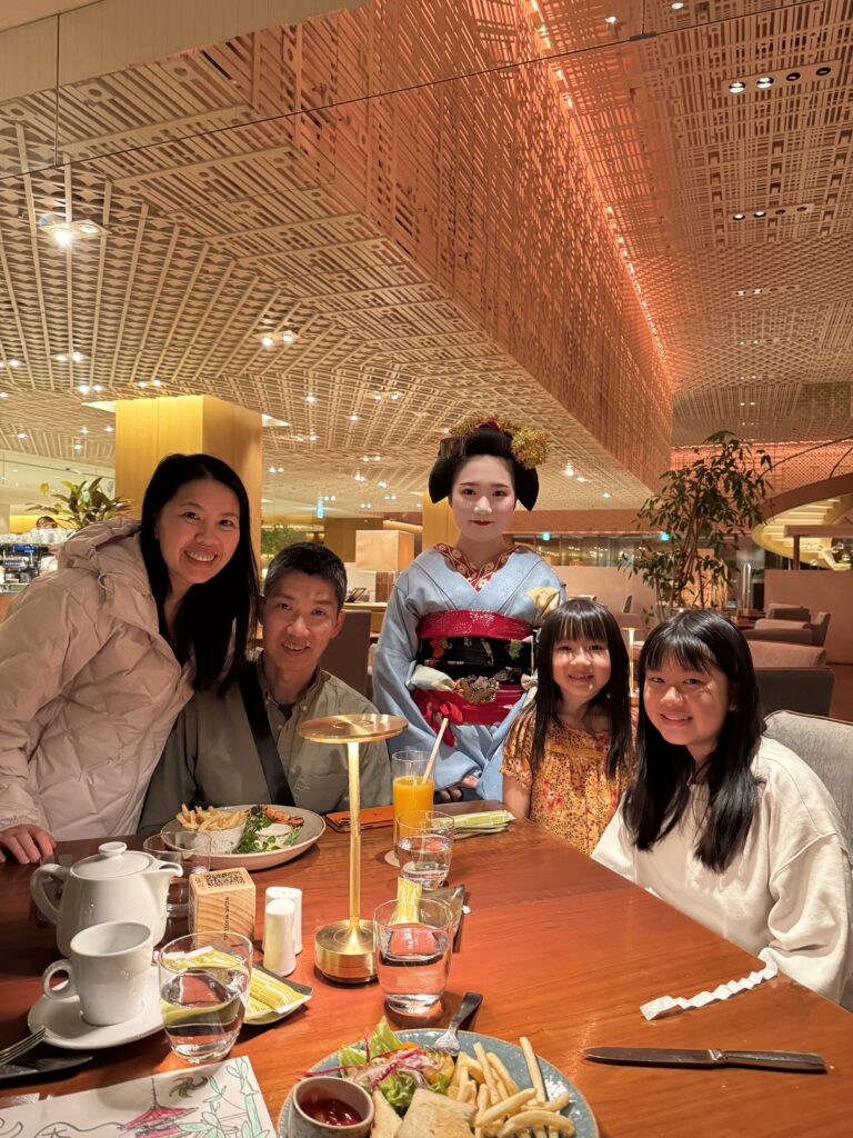 A family of four poses at a restaurant table with a woman dressed as a geisha. Plates of food and drinks are on the wooden table, and the background features warm lighting and a modern interior design.