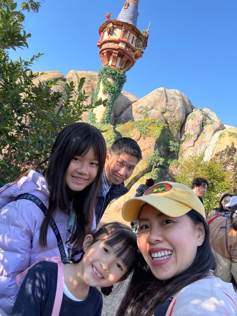 A smiling family of four poses for a selfie outside, with a tall, whimsical tower covered in ivy and rocks in the background under a clear blue sky.