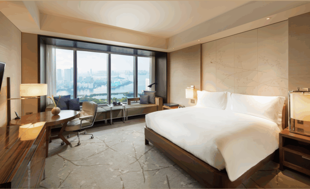 A modern hotel room with a large bed, desk, armchair, and window seat overlooking a cityscape and river. The room is well-lit and neutrally decorated, an ideal spot to relax or learn how to use rewards points for your next stay.