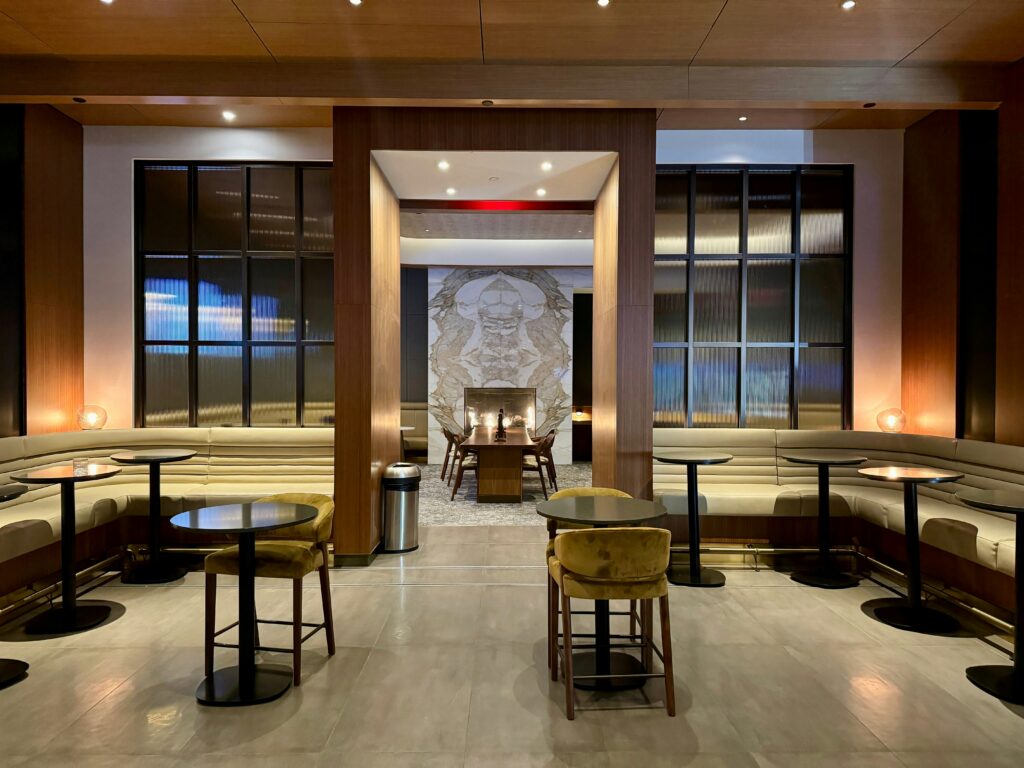 A modern, stylish lounge with beige cushioned seating, round tables, and velvet chairs. A large wall art design is visible at the back, framed by wooden panels and soft, warm lighting throughout the space.
