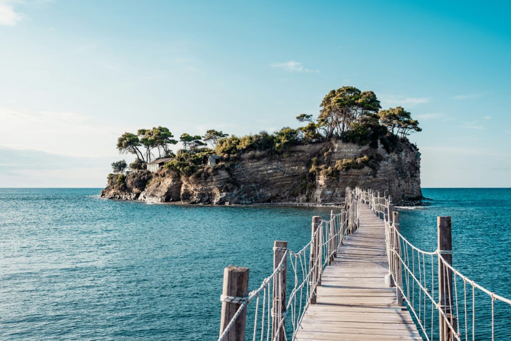 A wooden footbridge with rope railings leads over blue-green water to a small, rocky island covered with trees and shrubs under a clear blue sky.