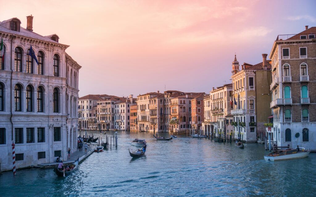 A scenic view of Venice’s Grand Canal at sunset, with historic buildings lining the water, gondolas and boats floating, and a pastel sky overhead.