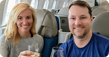 A woman and a man sit in airplane seats, smiling and holding up glasses, appearing to celebrate or enjoy a drink together during a flight.