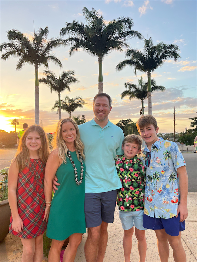 A family of five poses outdoors at sunset with palm trees in the background. The parents and three children smile, wearing summer clothes and colorful patterns. The sky is partly cloudy with a warm, golden glow.