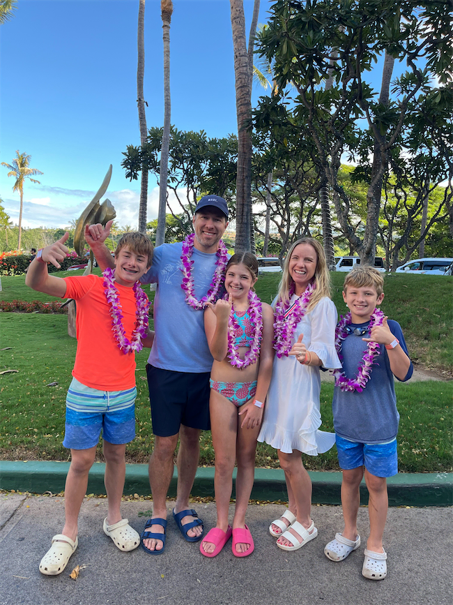A smiling family of five wearing leis poses outdoors on a sunny day. The parents and three children—two boys and a girl—stand together in casual summer clothes, with palm trees and green grass in the background.