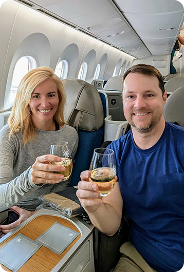 A smiling woman and man sit in airplane seats, holding glasses of white wine and toasting. They appear relaxed and happy in a bright, modern aircraft cabin.