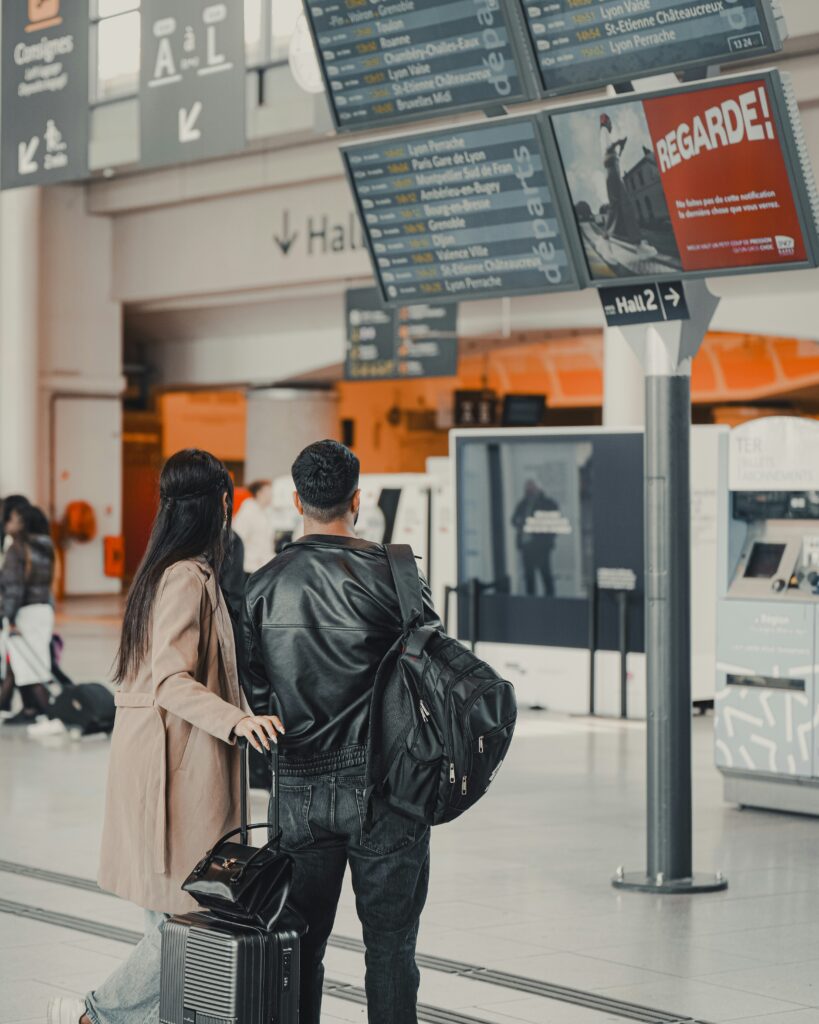 A man and woman stand close together in an airport terminal, looking up at a large departures board. They have luggage with them and appear to be checking flight information. Signs and schedules are visible above.